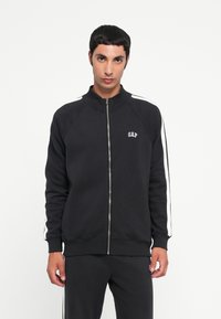 Felpa nera con zip e colletto alto, strisce bianche lungo le maniche e un piccolo logo "GAP" sul petto. Tessuto misto cotone.