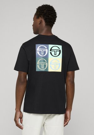 Uomo di spalle che indossa una maglietta nera con quattro loghi quadrati di diversi colori sulla schiena, abbinata a pantaloni bianchi.