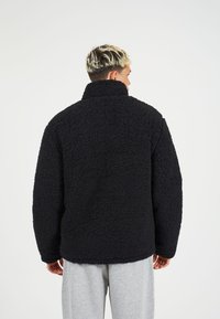 Juodas šalikas su tekstūru, pūsto paviršiaus, aukštu kaklu ir oversized stiliumi, dėvimas ant pilkų sportinių kelnių.