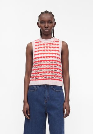 Donna con capelli intrecciati che indossa un top lavorato a maglia senza maniche rosa e rosso a motivi e jeans blu a vita alta, in piedi davanti a uno sfondo bianco semplice.
