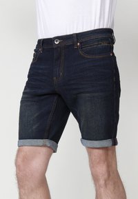 Pantalones cortos de denim en azul oscuro con dobladillo enrollado. Presenta un diseño clásico de cinco bolsillos y costuras en contraste. Fabricado con tela de algodón elástico.