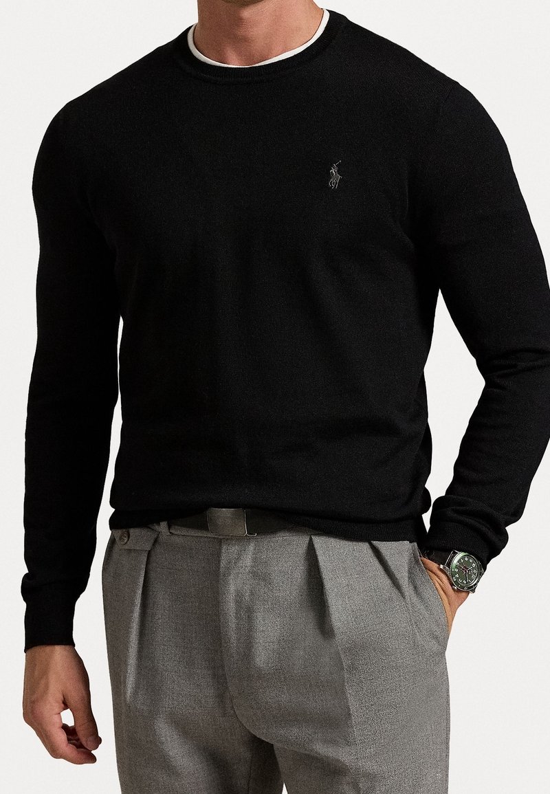 Homme portant un pull noir à manches longues avec un petit logo et un pantalon gris plissé, une main dans la poche et une montre visible au poignet.
