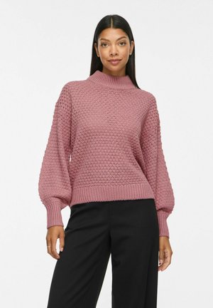 Femme portant un pull texturé mauve à manches longues avec col montant et un pantalon noir, debout et faisant face vers l'avant.