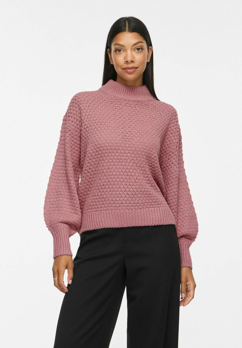 Femme portant un pull texturé mauve à manches longues avec col montant et un pantalon noir, debout et faisant face vers l'avant.
