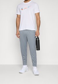 Camiseta blanca de manga corta con un logo de Nike multicolor, pantalones de chándal grises, zapatillas deportivas blancas y una botella de agua negra.