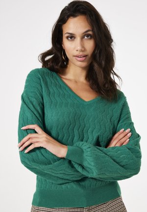 Pull en tricot vert avec un design en V, présentant un motif en zigzag texturé et des manches décontractées. Le tissu semble doux et confortable.