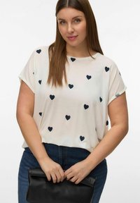 Vrouw met steil bruin haar, draagt een wit shirt met zwarte hartjes, blauwe spijkerbroek, en houdt een opgevouwen zwarte leren clutch vast.