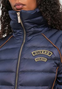 Mørkeblå puffjakke med høj krave, gylden lynlås og brune syninger. Har en tekstlogo-patch, der står "VIVERE CON STILE."