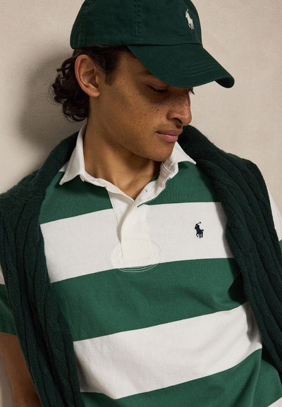 Jeune homme portant un polo rayé vert et blanc, une casquette vert foncé et un pull vert foncé posé sur les épaules, regardant vers le bas.