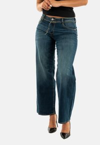 Jeans in denim a gamba larga di colore scuro con una leggera sbiaditura sulla parte anteriore. Presenta cinque tasche, chiusura con bottone e dettagli di cucitura discreti.