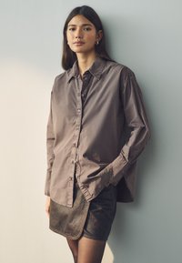 Long-sleeve, button-up shirt in muted brown fabric, paired with a dark brown leather mini skirt. Simple collar and relaxed fit.