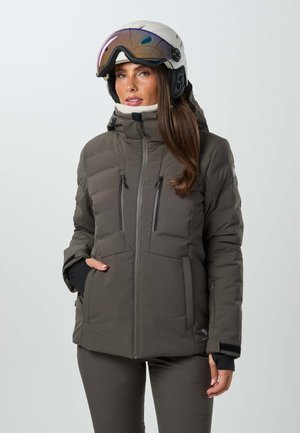 Femme portant une veste d'hiver matelassée vert foncé et un casque de ski blanc avec visière sur un fond uni.