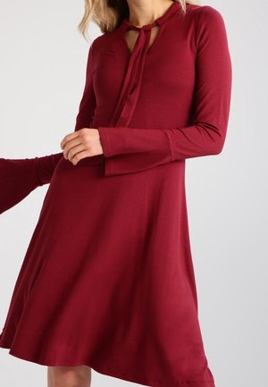 Femme portant une robe bordeaux à manches longues, longueur au genou, avec un col noué et une jupe évasée, tenant légèrement l'ourlet.