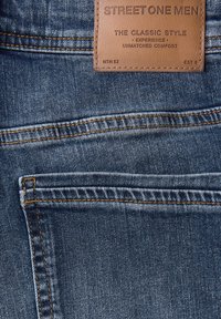 Poche arrière de jeans en denim bleu avec des coutures orange et une étiquette en cuir marron portant l'inscription "STREET ONE MEN Classic Style."