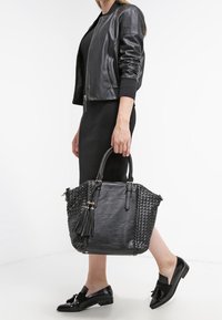 Svart läderhandväska med vävda sidor, handtag upptill och en tofsdetalj. Designen har en strukturerad form och en mjuk textur.