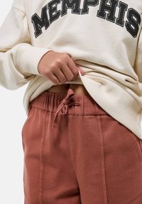 Personne soulevant un sweat-shirt crème avec le texte "MEMPHIS", portant un pantalon à cordon de couleur rouille, montrant une petite partie de son ventre.