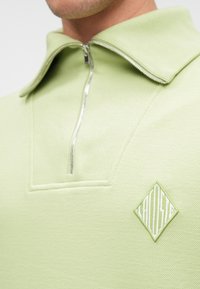 Ανοιχτό πράσινο πόλο με φερμουάρ και ραμμένο υφασμάτινο σχέδιο, διαθέτει λογότυπο σε σχήμα διαμαντιού σε ελαφρώς σκούρο πράσινο στο στήθος. Διαθέτει φαρδιά γιακά.