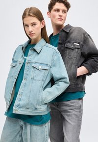 Νεαρή γυναίκα με ανοιχτόχρωμο τζιν μπουφάν και τζιν παντελόνι στέκεται μπροστά σε νεαρό άνδρα που φοράει μαύρο τζιν μπουφάν και γκρι τζιν παντελόνι.