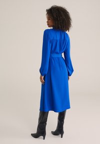 Rochie lungă albastră cu mâneci lungi, talie accentuată cu curea, textură moale și detalii pliate în spate; asortată cu cizme negre până la genunchi.