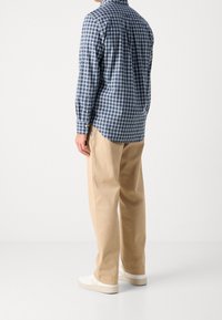 Mężczyzna ubrany w niebiesko-białą koszulę w kratę z długim rękawem, beżowe spodnie i białe trampki, stojący tyłem na białym tle.