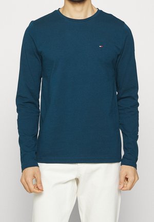 Homme portant une chemise bleue à manches longues avec un petit logo sur la poitrine et un pantalon blanc, debout devant un fond uni.
