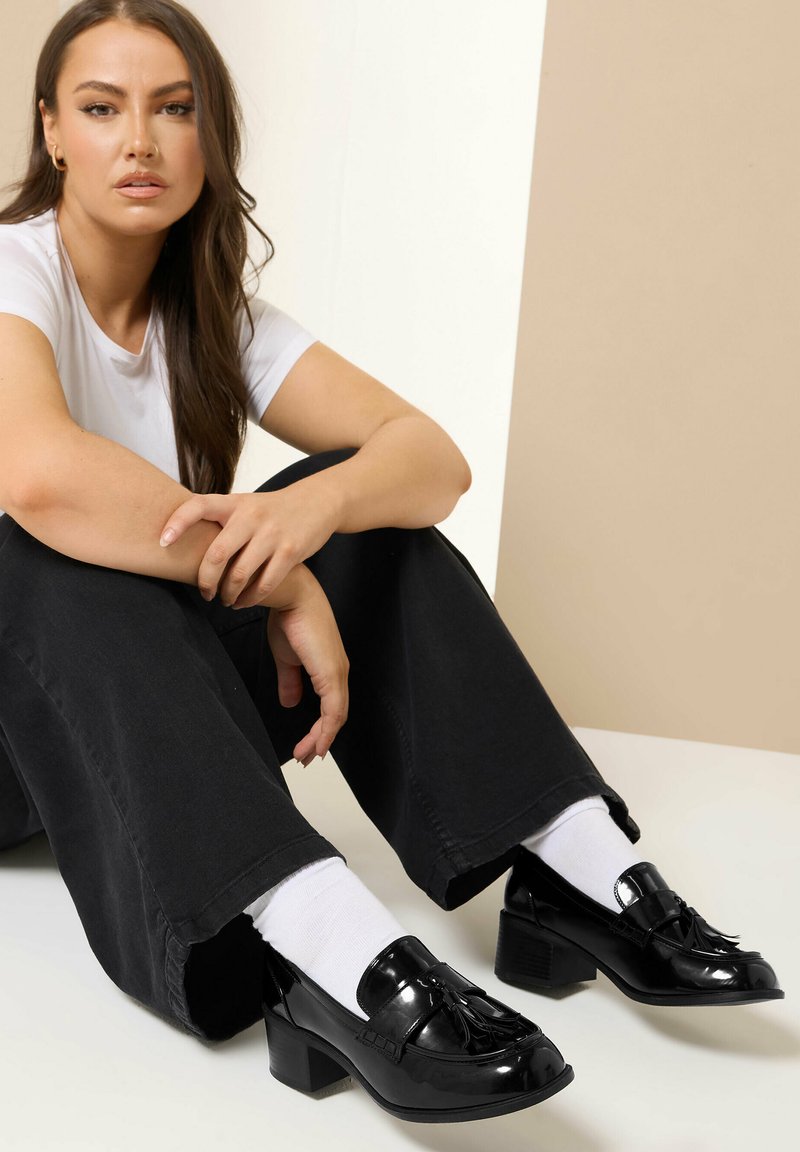 Svarta lackläderskor med blockklack och detaljer i fransar, som bärs med vita strumpor och lösa svarta byxor, sittande mot en neutral bakgrund.