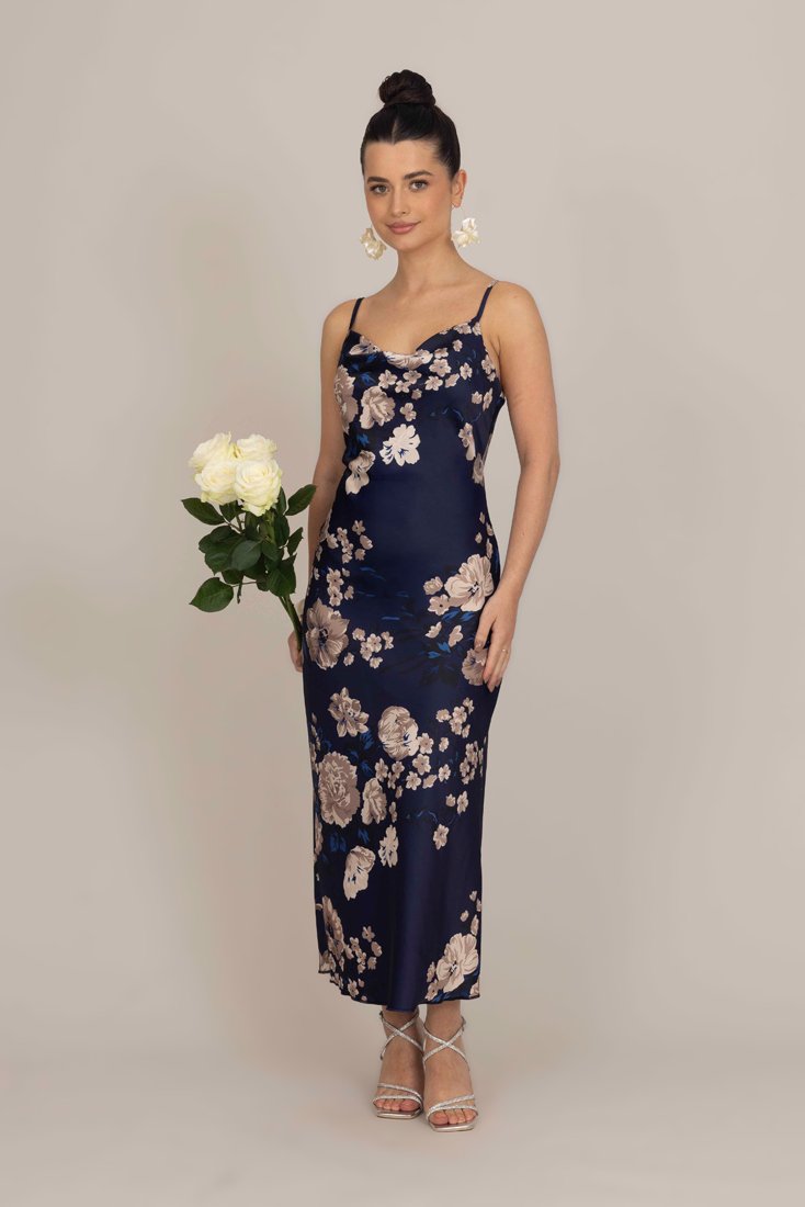 Abito in satin blu navy floreale con spalline sottili e una silhouette aderente. Presenta grandi fiori beige e dettagli blu. Il modello tiene in mano rose bianche.