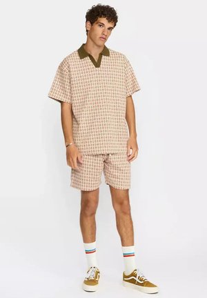 Joven de pie, vestido con camisa de manga corta y pantalones cortos beige con estampado a juego, calcetines blancos con rayas de colores y zapatillas marrones.