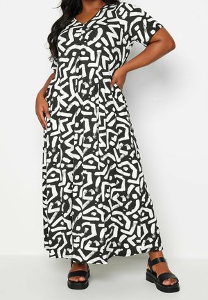 Femme portant une robe maxi à manches courtes avec un motif abstrait noir et blanc et des sandales à plateforme noires, debout d'une main sur la hanche.