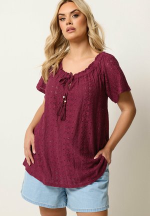 Kobieta ubrana w teksturowaną bordową bluzkę z krótkim rękawem i sznurowaniem z chwostami oraz jasnoniebieskie krótkie spodenki dżinsowe, stoi z rękami w kieszeniach.