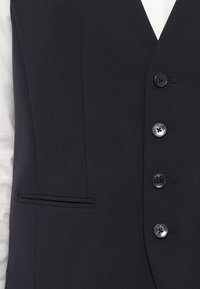 Tamsiai pilkas liemenė, pagaminta iš lygaus audinio. Yra V formos kaklo iškirptė, penki juodi mygtukai ir šoninė kišenė su elegantišku dizainu.