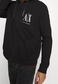 Felpa nera con zip realizzata in tessuto morbido. Presenta un logo bianco sul fronte, cappuccio con cordino e polsini e orlo a coste.