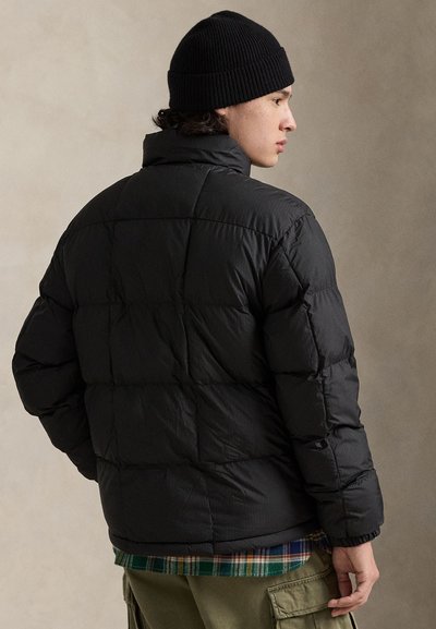 Juoda pūsta striukė su siuvinėjimu, aukšta apykakle ir elastingais rankogaliais, dėvima ant žalios languotos marškinės ir alyvinių kario stiliaus kelnių.