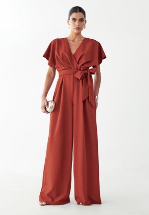 Žena oblečená v jumpsuite s širokými nohavicami hrdzavej farby, s výstrihom do V, krátkymi zvonovými rukávmi, opaskom viazaným v páse, držiaca malú kovovú kabelku.