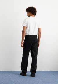 Μαύρο cargo παντελόνι με πολλές τσέπες, φτιαγμένο από ανθεκτικό ύφασμα. Συνδυασμένο με ένα απλό λευκό μπλουζάκι, παρουσιάζεται από πίσω.