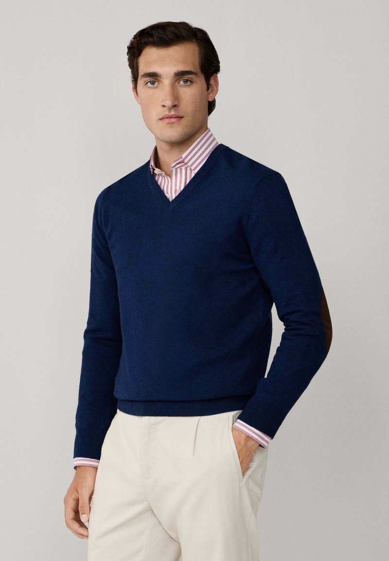 Suéter de punto azul marino con cuello en V, que presenta parches marrones en los codos. Combinado con una camisa de color rosa y blanca a rayas con cuello debajo.