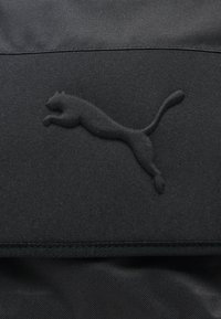 Ανάγλυφο μαύρο λογότυπο Puma με μια γάτα που πηδά σε υφή υφάσματος σκούρου χρώματος, πιθανώς μέρος τσάντας ή ρούχου.