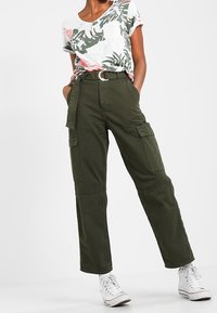 Oliivinvihreät cargo-housut, joissa on isot sivutaskut, leveät lahkeet ja yhteensopiva vyö, yhdistettynä lyhythihaiseen trooppikuvioiseen paitaan.