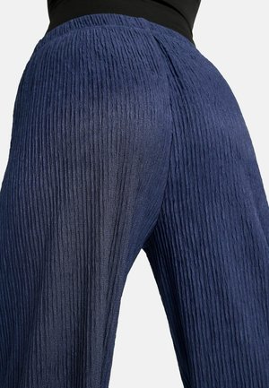 Žena nosící tmavě modré široké kalhoty s vysokým pasem a texturou a černý top, zobrazená od pasu dolů na světle pozadí.