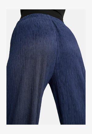 Mujer que lleva pantalones anchos de talle alto, texturizados, de color azul marino, y una blusa negra, mostrada desde la cintura hacia abajo sobre un fondo claro.