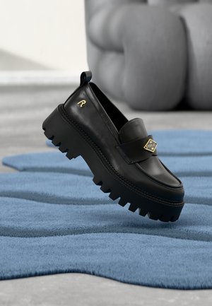 Musta nahkainen loafer-kengän paksulla, uritetulla pohjalla. Koristeena kultainen logoyksityiskohta ja päällä oleva remmi. Aseteltu sinisen tekstuuripintaisen maton päälle.