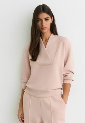 Hosszú sötét hajú nő pasztell rózsaszín pulóver pulóverben és hozzáillő, bő nadrágban, egyik kezét zsebébe süllyesztve, fehér falnak támaszkodva áll.