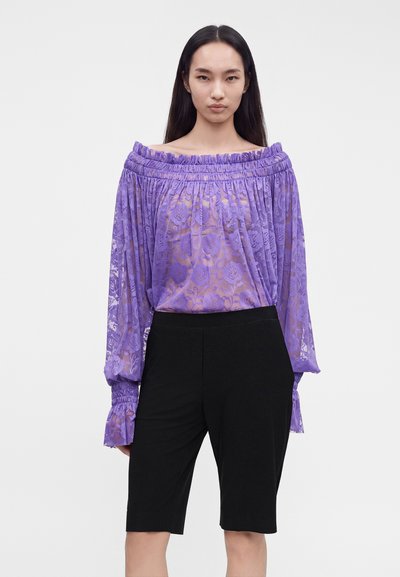 Femme aux longs cheveux foncés portant un chemisier en dentelle violet transparent à épaules dénudées et un short noir à la hauteur des genoux, debout devant un fond blanc.