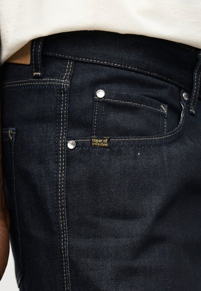 Tumesiniste teksapükste tasku lähivõte hõbedaste naeltega ja väike "tiger of sweden" silt vöö all ning valge särk peal.