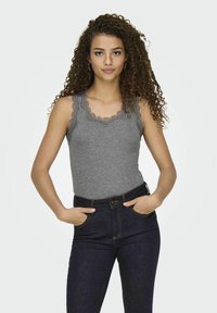 Grå tanktop med blondekant ved halsudskæringen, parret med mørkeblå højtaljede jeans. Modellen står med hænderne i lommerne.