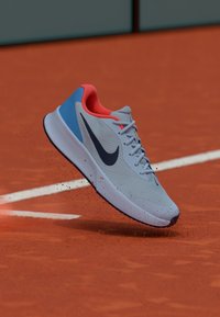 Šviesiai pilka sportinė avalynė su mėlyna akcentu, oranžine apykakle ir juodu „swoosh“ ženklu. Viršus turi tekstūruotą dangą, o baltas guminis padas pakeltas virš molio korto.