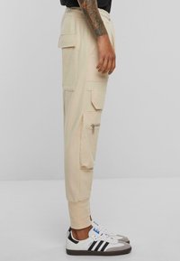 Beige cargo bukser med sidelommer, lynlåsdetalje og ribbede ankler, parret med hvide sneakers med sorte og grå accenter.