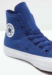 Blue canvas high-top sneaker with white rubber sole. Features eyelets, textured fabric, and a circular logo patch on the side.
