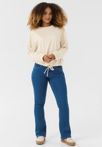 Maglietta beige a maniche lunghe con orlo elastico, abbinata a jeans a zampa d'elefante in denim blu e sneakers beige con dettagli rossi. Tessuto morbido, vestibilità casual.