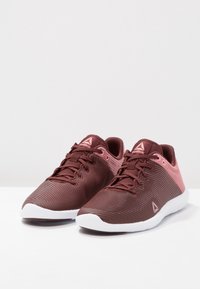 Bordeauxrode en roze sportschoenen met textiel, witte zolen en een vetersluiting. De schoenen hebben een logo op de tong en aan de zijkant.
